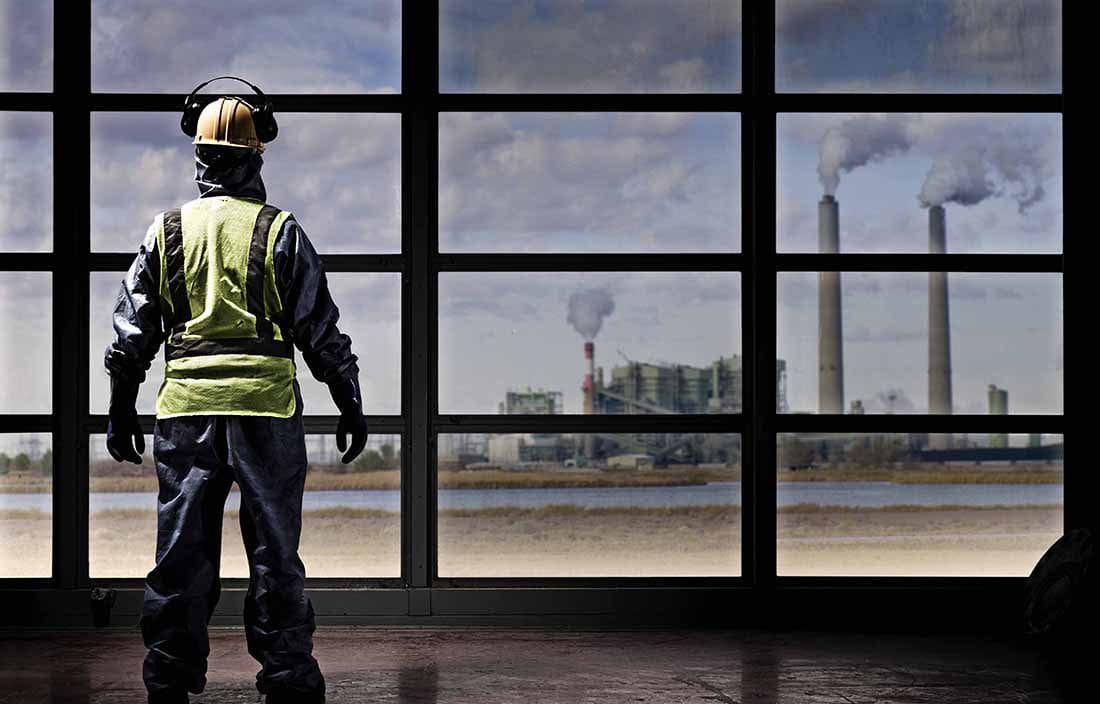 Construction worker looking out a window towards an industrial factory site.
